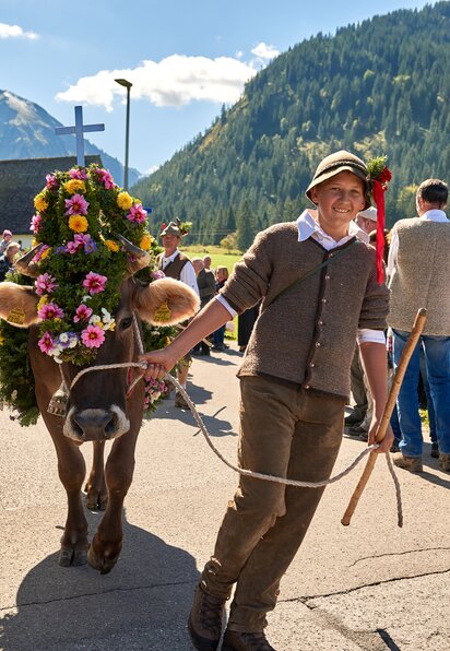 Mann führt geschmückte Kuh beim Almabtrieb durch ein Dorf, im Hintergrund Kirche und Berge