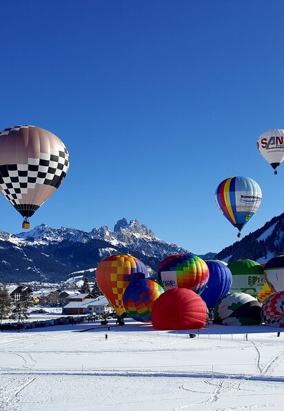 Ballonfestival im Winter mit vielen bunten Heißluftballons vor schneebedeckten Bergen unter blauem Himmel