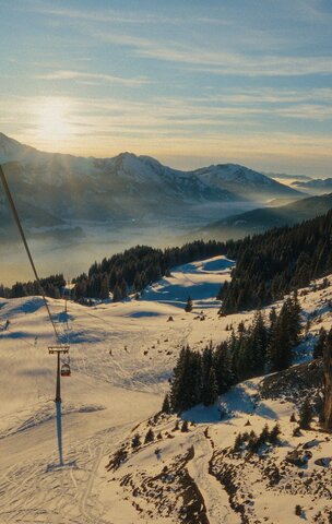 Winterlandschaft mit schneebedeckten Bergen, Skipiste, Sessellift und bewaldeten Hängen im Sonnenlicht