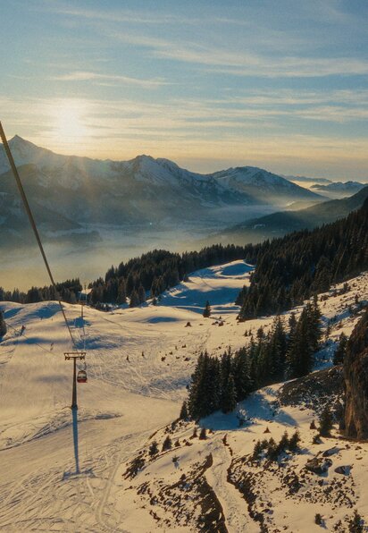 Winterlandschaft mit schneebedeckten Bergen, Skipiste, Sessellift und bewaldeten Hängen im Sonnenlicht