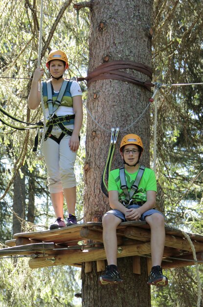 Zwei Kinder mit Helmen und Klettergurten auf einer Plattform im Kletterwald