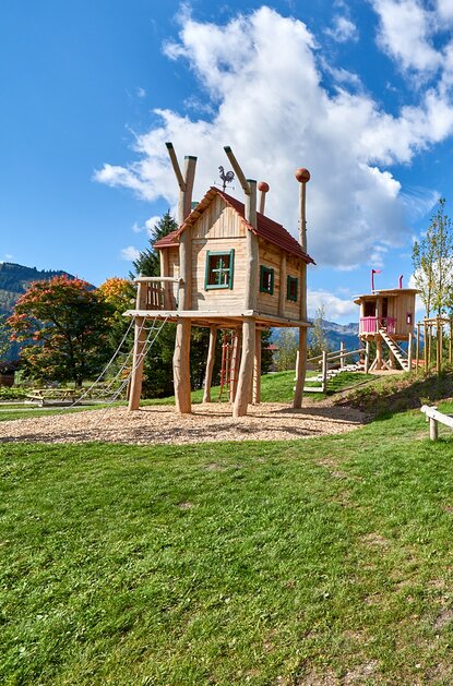Spielplatz mit Holzhäusern, Rutsche und Klettergerüsten auf grüner Wiese vor Wald und blauem Himmel