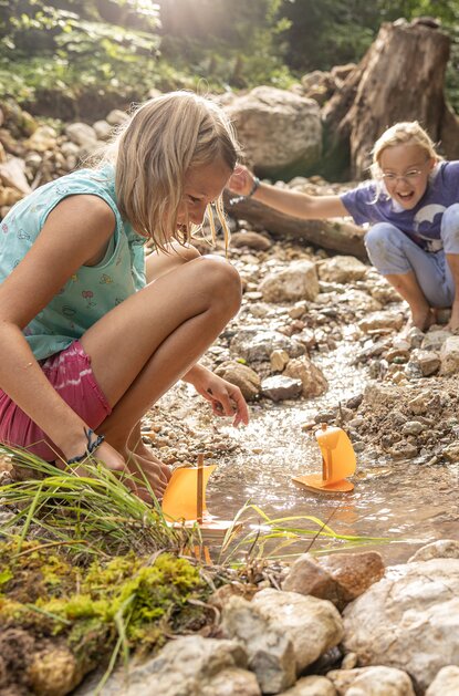 Zwei Mädchen spielen mit kleinen Segelbooten in einem Bachbett