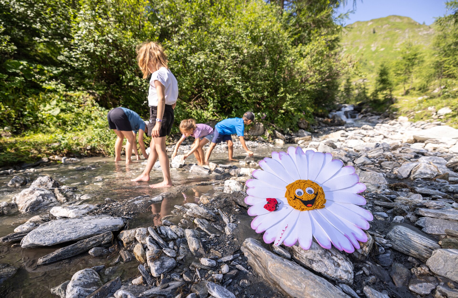 Kinder spielen barfuß in einem Bach, eine große Stoffblume liegt auf Steinen