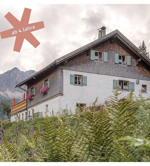 Haus mit Holzfassade und grünen Fensterläden vor Bergkulisse, Hinweis "ab 4 Jahre"