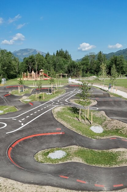 Pumptrack-Anlage mit Hügeln und Kurven in Parklandschaft vor Bergkulisse