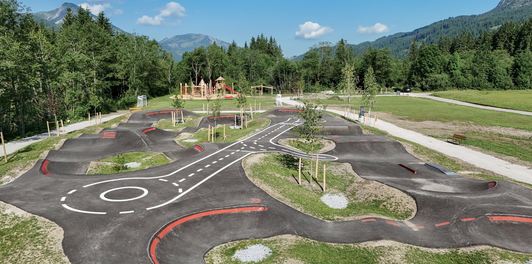 Pumptrack-Anlage mit Hügeln und Kurven in Parklandschaft vor Bergkulisse