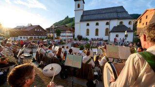 Blasorchester spielt vor weißer Kirche im Freien, Publikum sitzt auf Bänken