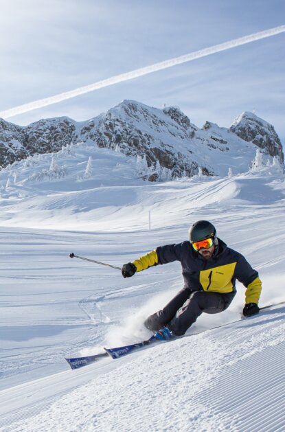 Skifahrer in gelb-schwarzer Jacke fährt vor schneebedecktem Berg
