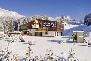 Schneebedeckte Skischule mit Schneemann-Motiv vor Berglandschaft an sonnigem Wintertag
