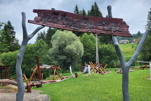 Holzschild "Märchenpfad" vor Spielplatz mit Baumstämmen und grüner Wiese