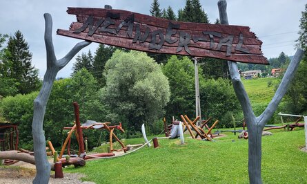 Holzschild "Märchenpfad" vor Spielplatz mit Baumstämmen und grüner Wiese