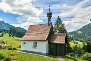 Kleine Kapelle mit braunem Dach auf grüner Wiese vor bewaldeten Hügeln unter blauem Himmel mit Wolken