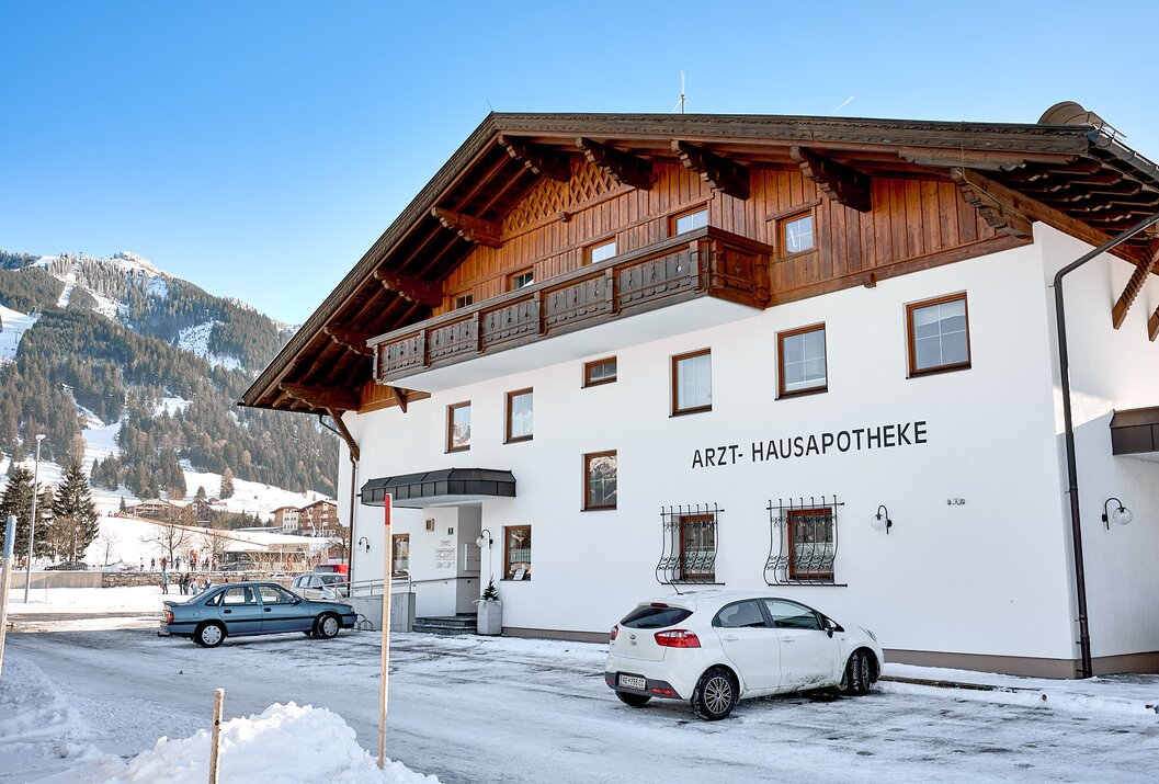 Weißes Gebäude mit Aufschrift "Arzt-Hausapotheke" und zwei geparkten Autos im Winter