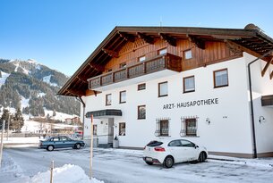 Weißes Gebäude mit Aufschrift "Arzt-Hausapotheke" und zwei geparkten Autos im Winter
