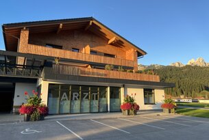Arztpraxis in Holzhaus mit Parkplatz und Blumenkästen vor Panorama