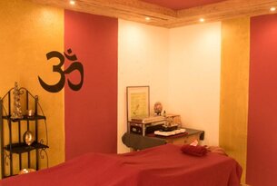 Massageraum mit roter Liege, Regal, Om-Symbol an gelb-roter Wand und Holzdecke