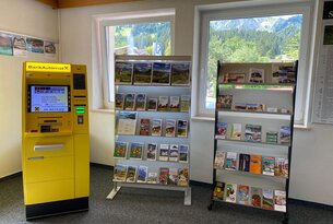 Gelber Automat und zwei Regale mit Broschüren vor Fenstern