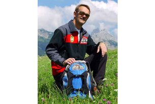 Mann kniet mit Rucksack auf grüner Wiese vor Bergpanorama