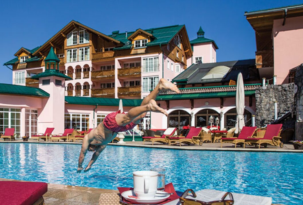 Mann springt in Pool vor Hotelanlage mit Liegen, Tisch, Tasse, Zeitung und Sonnenbrille