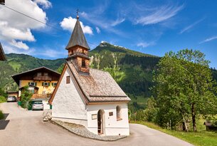 Weiße Kapelle mit dunklem Spitzdach vor Berglandschaft und blauem Himmel