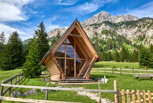 Holzkapelle vor Bergkulisse auf grüner Wiese
