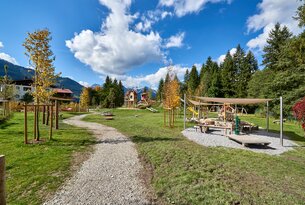 Spielplatz mit Holzhäusern, Sandkasten und Schaukeln vor Bergkulisse unter blauem Himmel mit Wolken