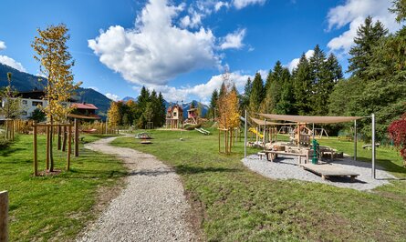 Spielplatz mit Holzhäusern, Sandkasten und Schaukeln vor Bergkulisse unter blauem Himmel mit Wolken
