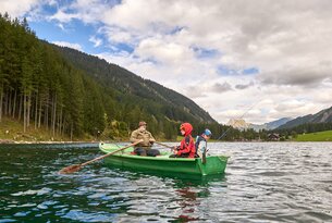 Mann und zwei Kinder angeln in einem grünen Ruderboot auf einem See vor bewaldeten Bergen