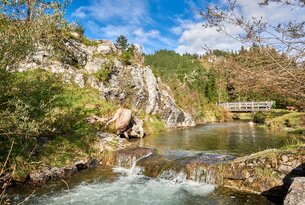 Flusslauf mit kleinem Wasserfall, Felsen, Bäumen und Brücke vor bewaldetem Hügel unter blauem Himmel