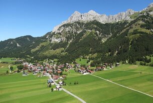 Dorf in grüner Landschaft vor Bergmassiv unter blauem Himmel