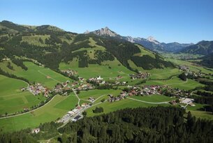 Luftaufnahme eines Dorfes in grüner Hügellandschaft vor Bergpanorama