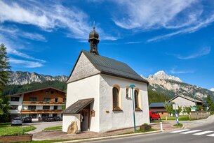 Kleine weiße Kapelle mit Glockenturm vor Bergkulisse unter blauem Himmel