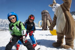 Zwei Kinder mit Skiausrüstung im Schnee, Maskottchen und Mammut-Skulptur im Hintergrund