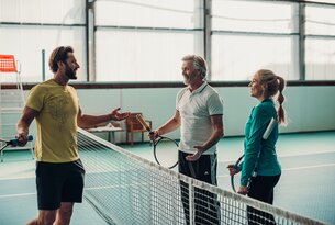 Drei Personen mit Tennisschlägern unterhalten sich am Netz auf einem Tennisplatz