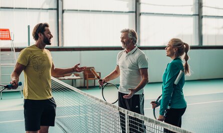 Drei Personen mit Tennisschlägern unterhalten sich am Netz auf einem Tennisplatz
