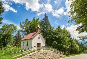 Kleine weiße Kapelle mit rotem Dach vor Bäumen und blauem Himmel mit Wolken