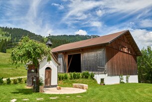 Holzscheune neben kleiner Kapelle mit Baum vor Bergkulisse