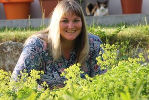 Frau lächelt im Garten, umgeben von Pflanzen, im Hintergrund Blumentöpfe und eine Katze im Katzenhaus