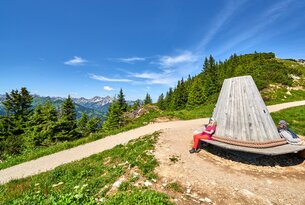 Frau sitzt auf Bank mit Bergblick an Wanderweg im Grünen unter blauem Himmel