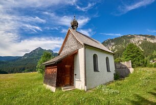 Kleine weiße Kapelle mit Holzdach vor Bergkulisse und blauem Himmel