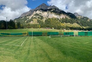 Grüner Sportplatz mit Netzen vor Berglandschaft mit bewaldeten Hängen und felsigem Gipfel