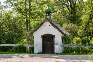 Kleine weiße Kapelle mit grünem Turm und Holztür vor Bäumen