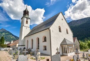 Weiße Kirche mit Turm und dunklem Dach vor blauem Himmel und Bergen