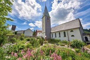 Kirche mit hohem Turm, umgeben von Blumen und Häusern unter blauem Himmel mit Wolken
