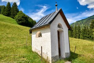 Kleine weiße Kapelle auf grüner Wiese vor bewaldetem Hang unter blauem Himmel