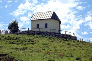 Kleine Kapelle auf einem Hügel mit Holzzaun und Steinmauer vor blauem Himmel