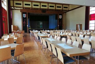 Leerer Saal mit Tischen und Stühlen vor einer Bühne mit blauem Vorhang
