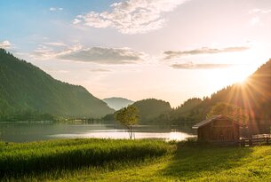 See mit Bergen im Hintergrund, Wiese im Vordergrund und Hütte am rechten Bildrand