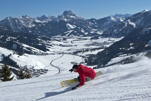 Snowboarder in roter Jacke fährt Hang hinab, verschneite Berge im Hintergrund